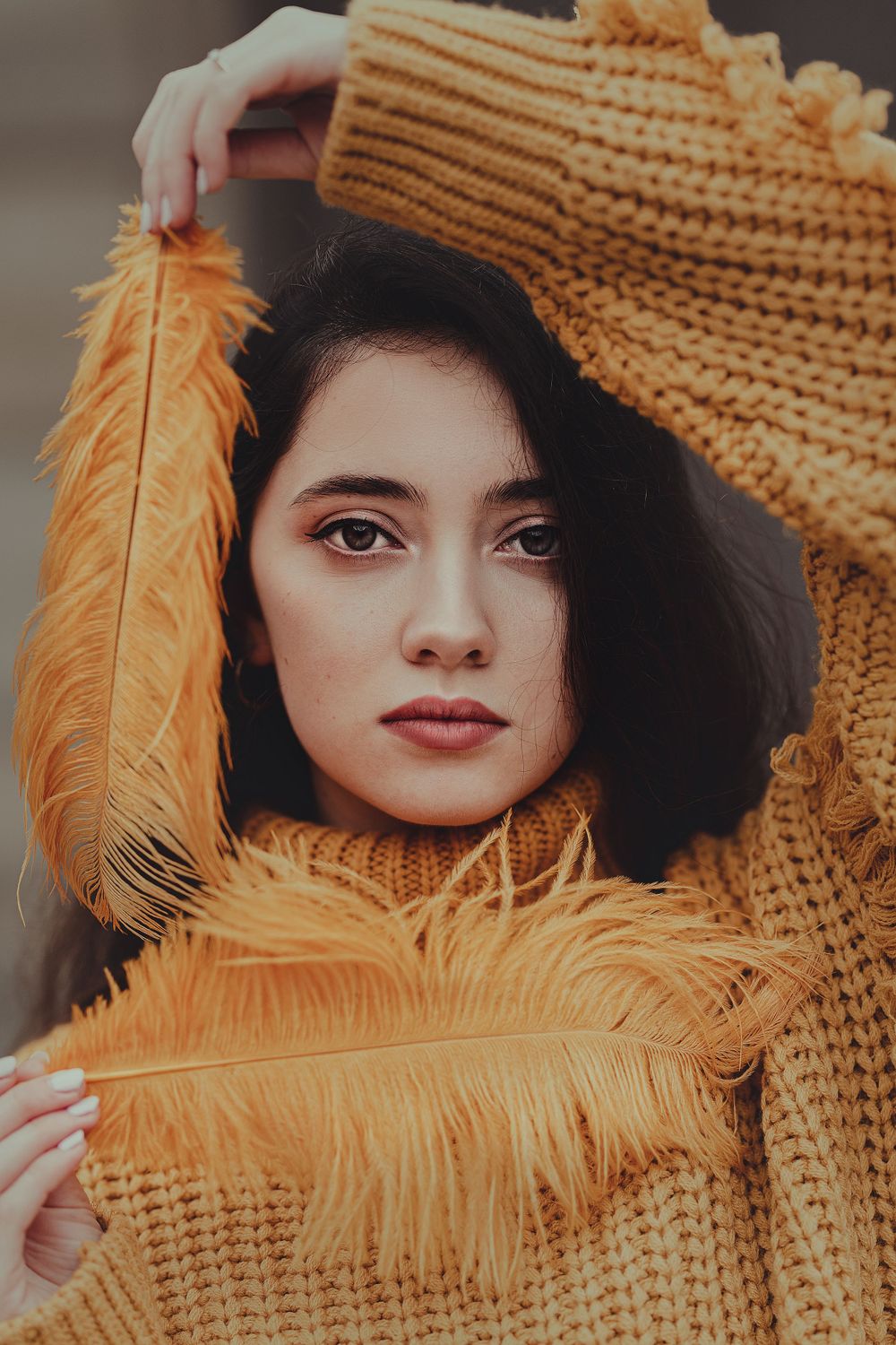 Artistic portrait with orange feather accent against mustard sweater and black hat.