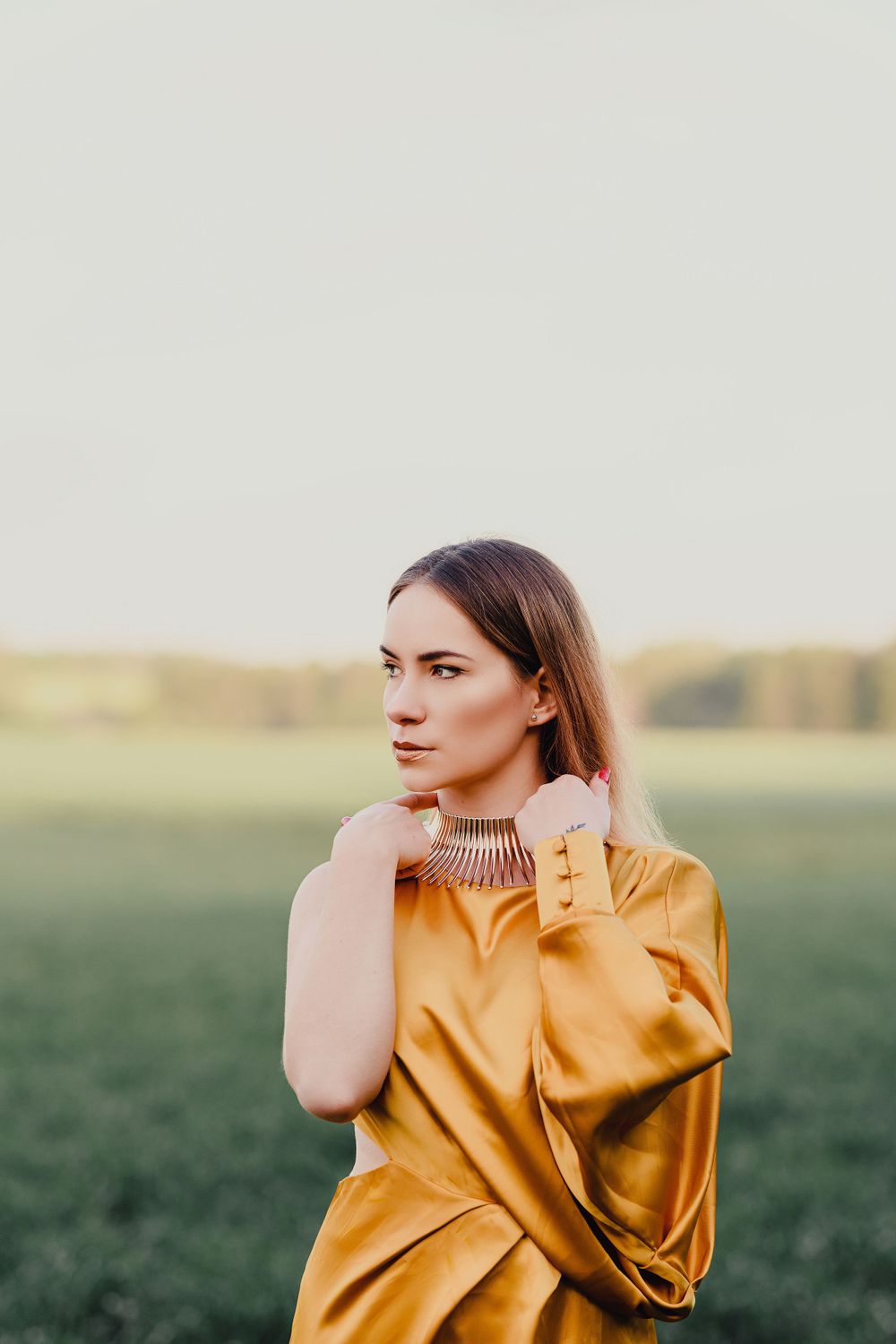 Stylish portrait in golden dress with statement necklace against natural outdoor backdrop.