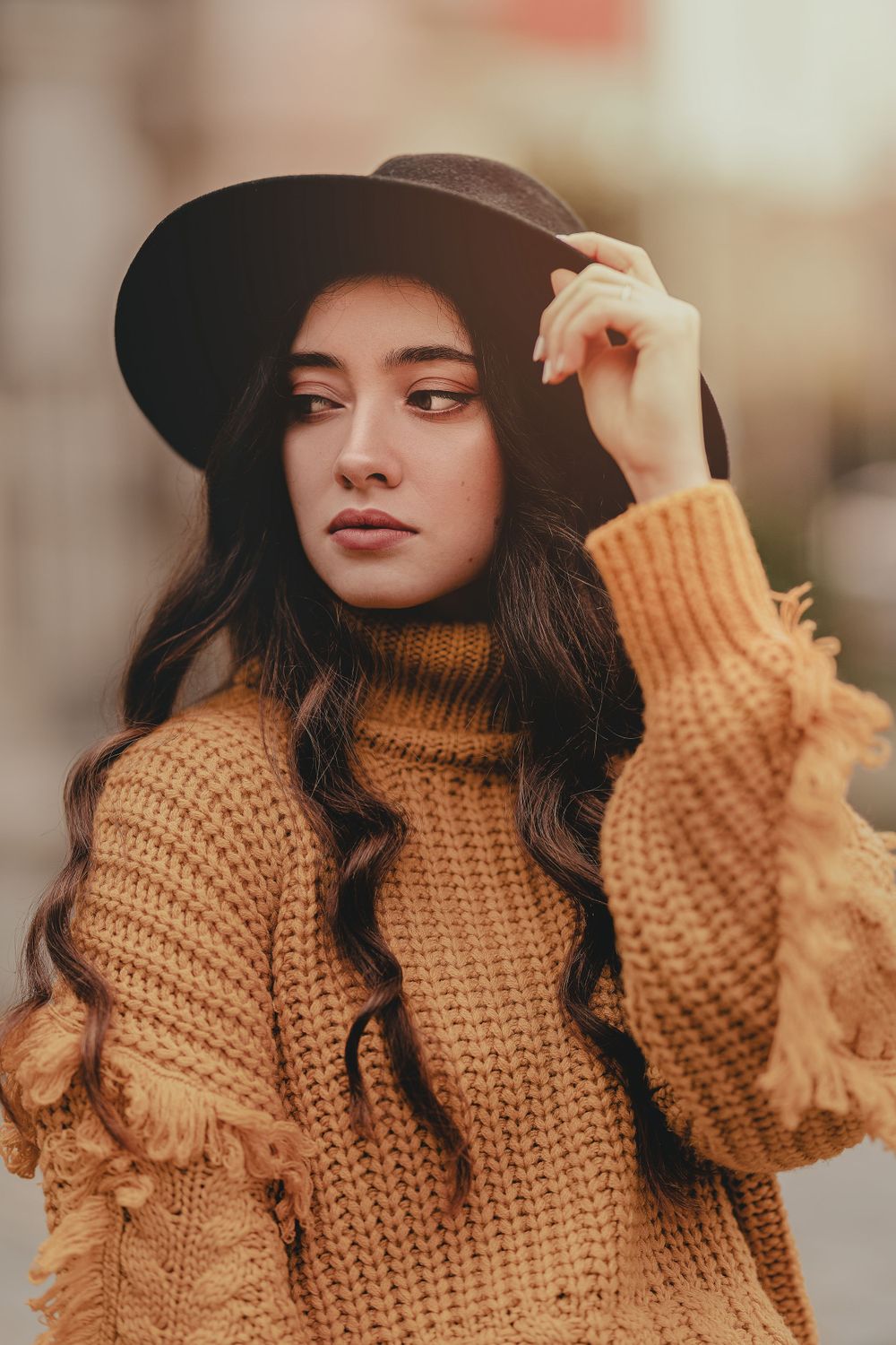 Fashion portrait featuring a chunky knit mustard sweater and black wide-brim hat against a warm autumn background.