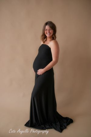 Maternity photo session flowing black strapless gown poses elegantly against a neutral brown backdrop in a studio setting. Hartford, CT