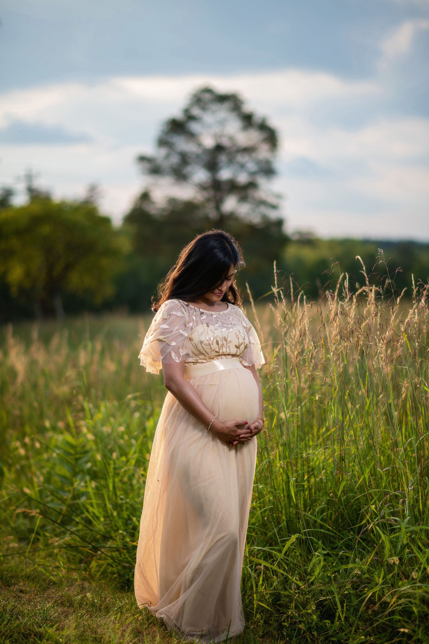 Woman in flowing cream dress walks through tall grass field at sunset.