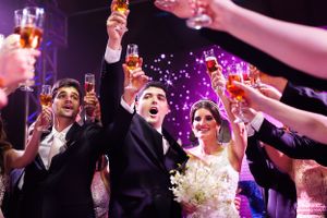 Wedding reception guests raise glasses in celebration under purple party lights.