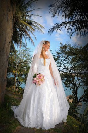 A bride in a flowing white dress holds a pink bouquet while standing among palm trees by the ocean.