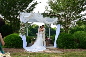 Couple shares intimate moment under white draped pergola during garden wedding ceremony.