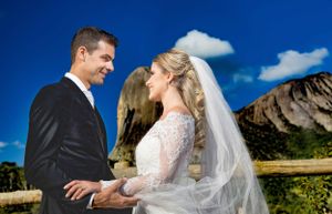 A wedding couple poses against a dramatic mountain backdrop with blue skies and white clouds.