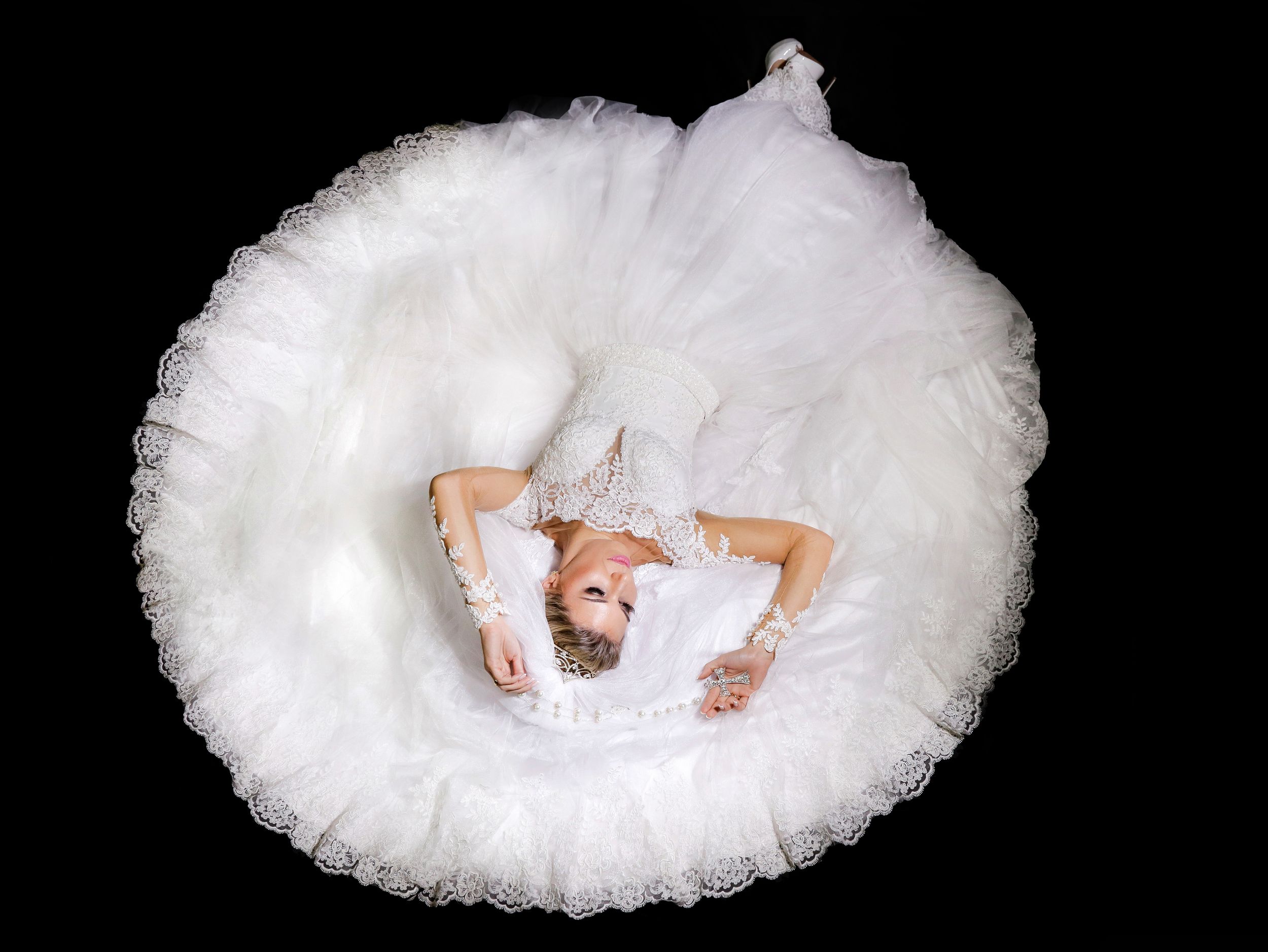 An overhead view of a bride in a circular white wedding dress laying on a black background.