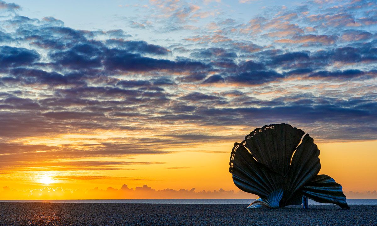 A large scallop shell silhouetted against a vibrant orange and blue sunset sky over the ocean.