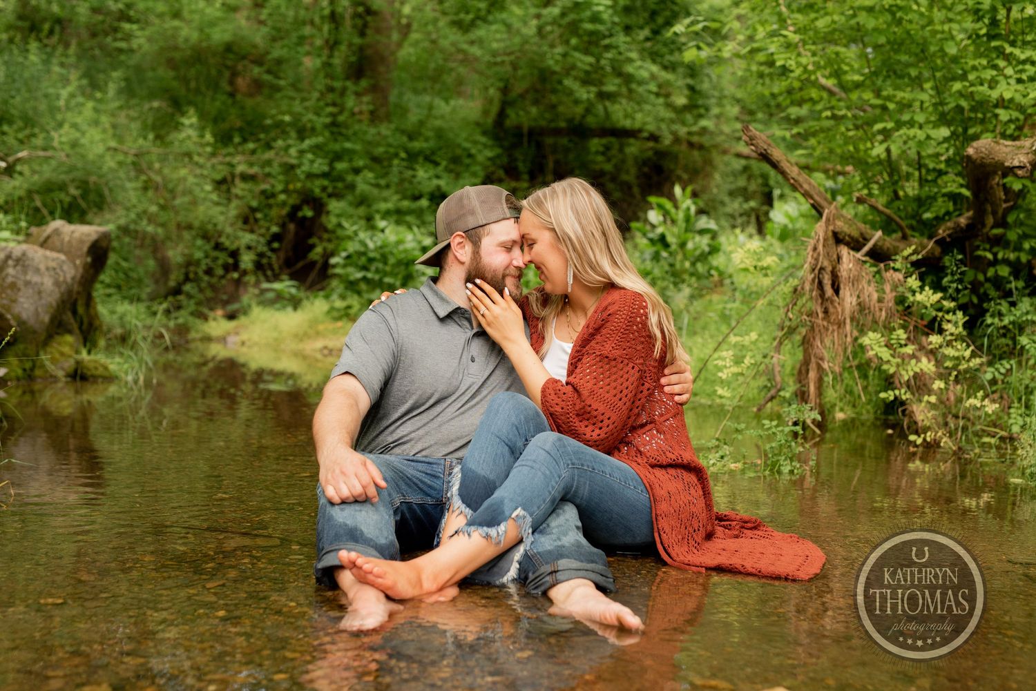 A couple laughs together while sitting in shallow creek waters surrounded by greenery.