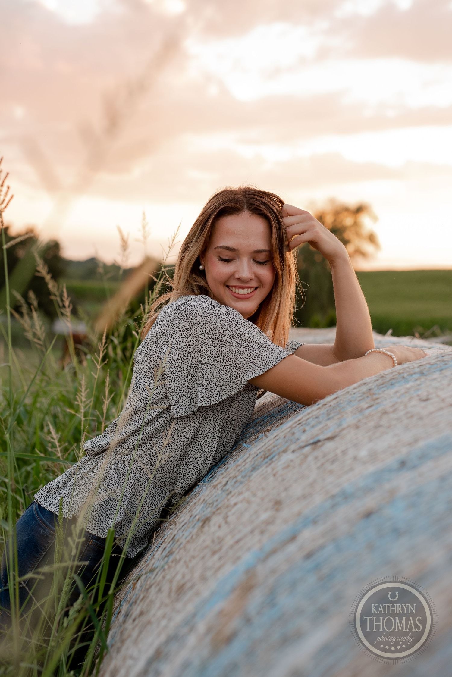 Casual outdoor portrait by a hay bale with soft sunset lighting in a rural field.