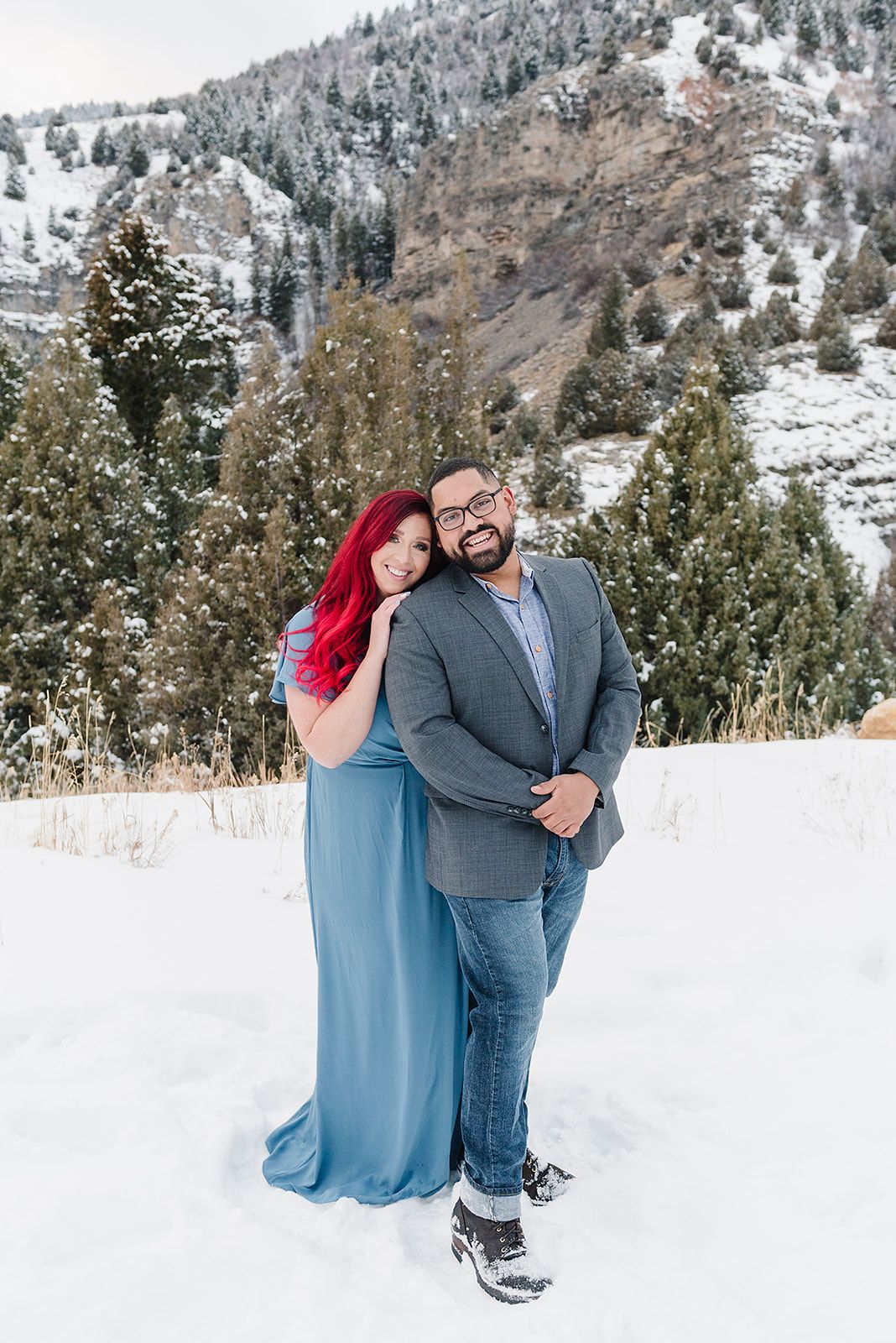 A couple poses together in a snowy mountain setting with evergreen trees behind them.