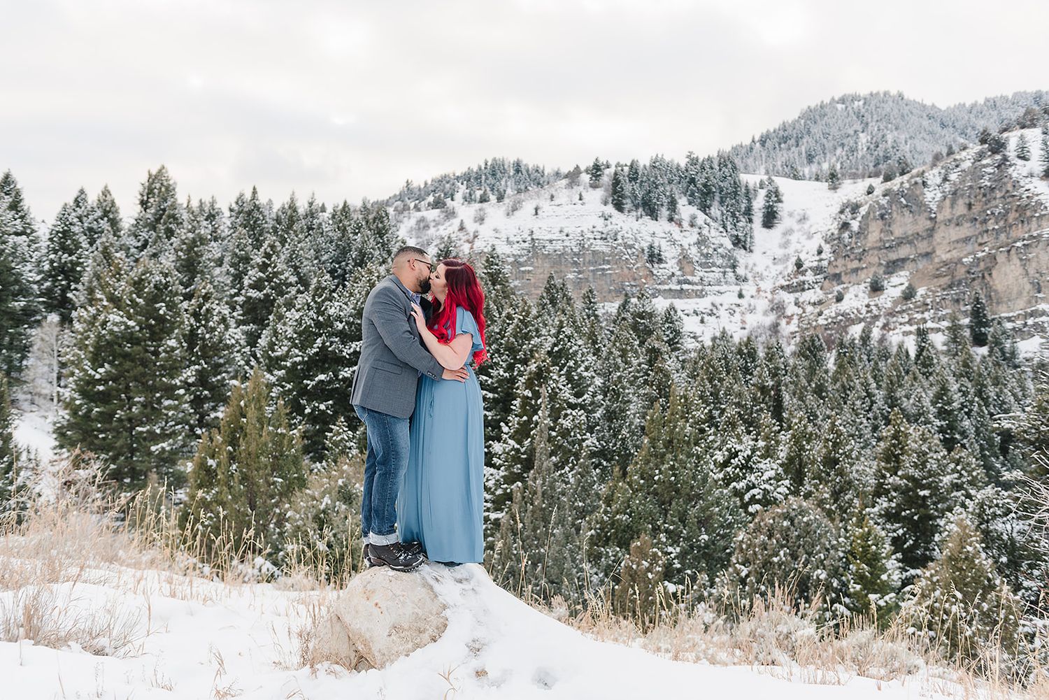 A couple embraces in a snowy mountain landscape surrounded by evergreen trees and winter scenery.