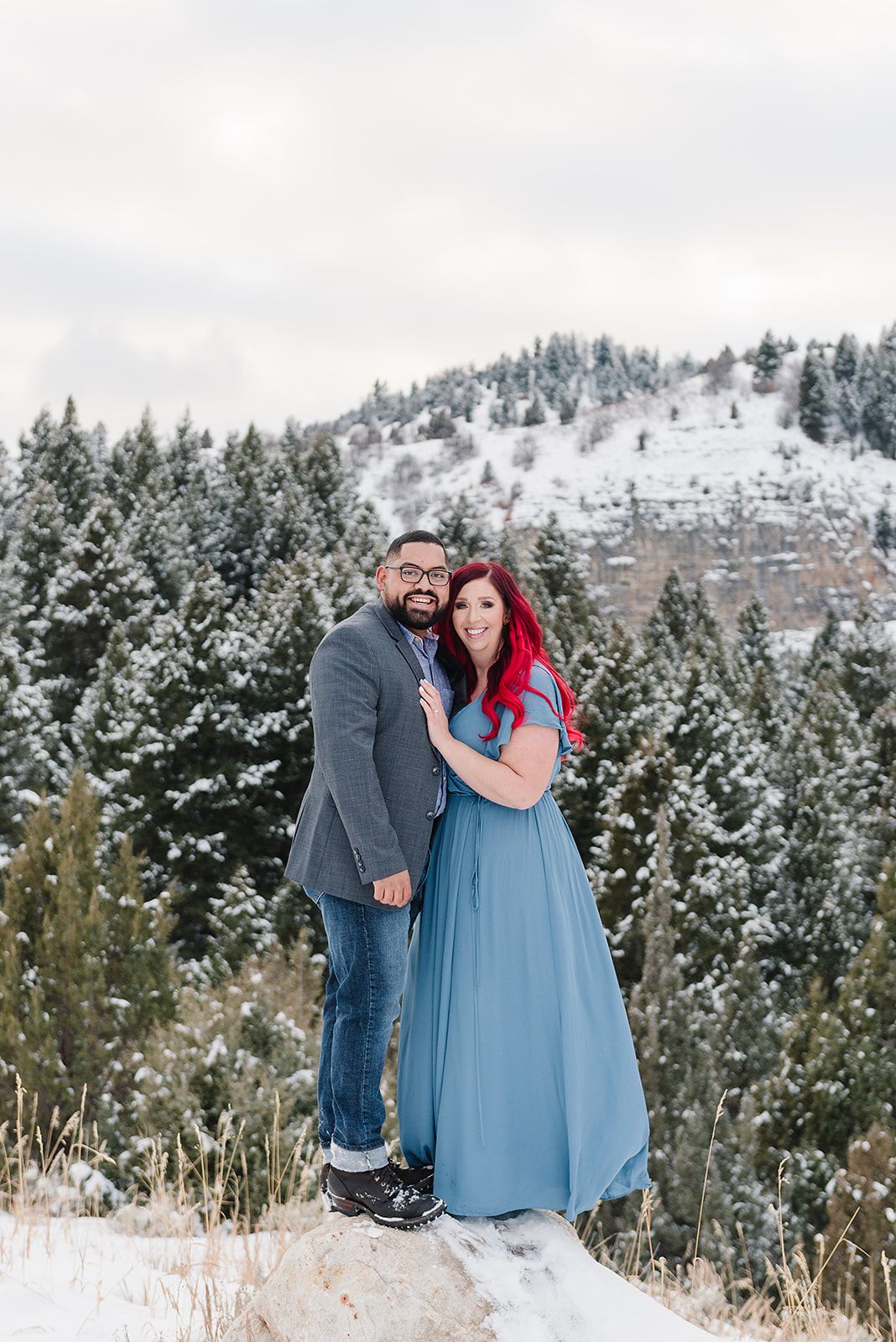 A couple poses together on a snowy rock outcropping with snow-covered pine trees and mountains in the background.