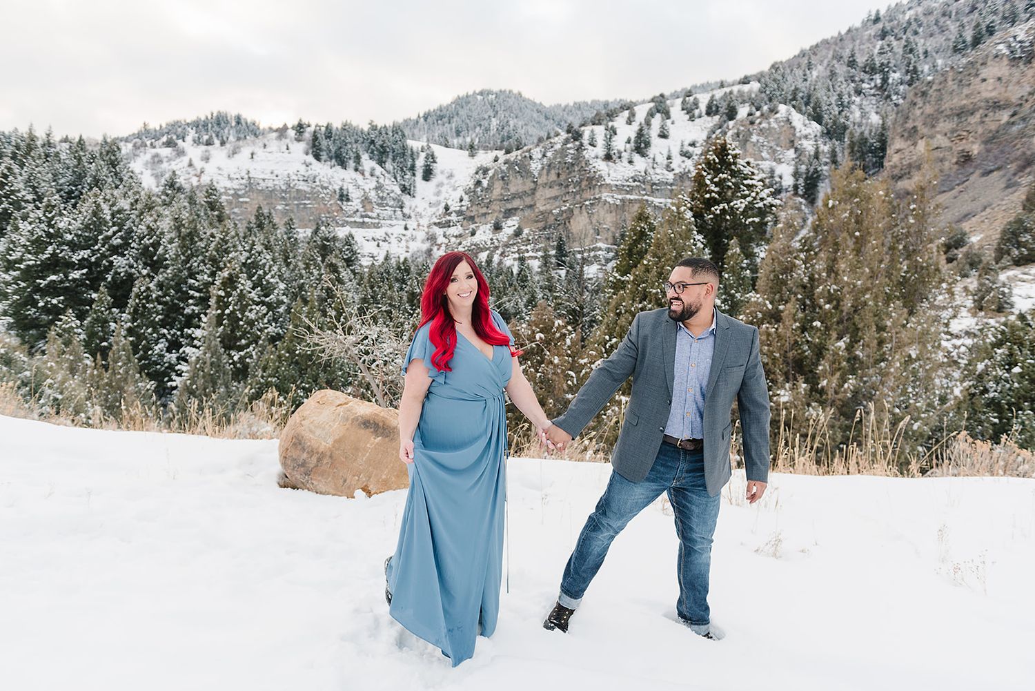 A couple holds hands while walking in a snowy mountain setting with evergreen trees.