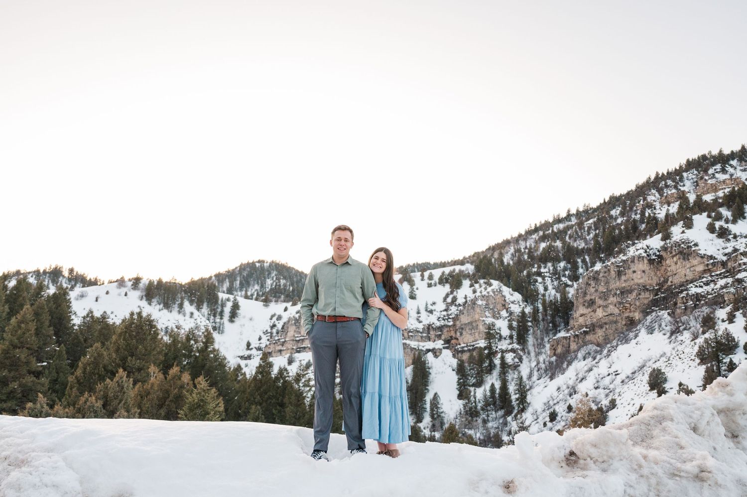 A couple walks together on a snowy mountain path with scenic winter landscape behind them.