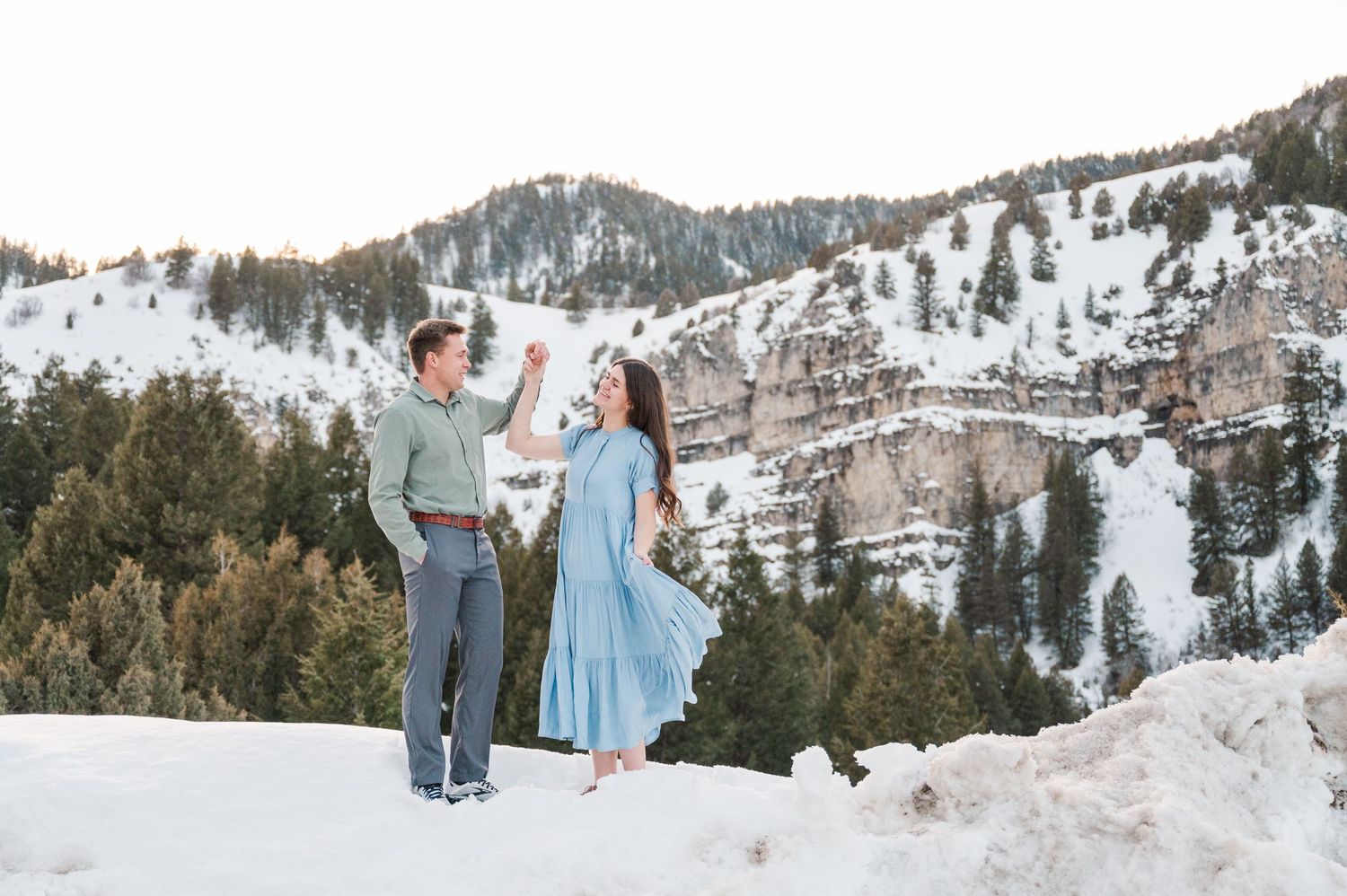 A couple dances in the snow with mountains and evergreen trees creating a winter wonderland backdrop.