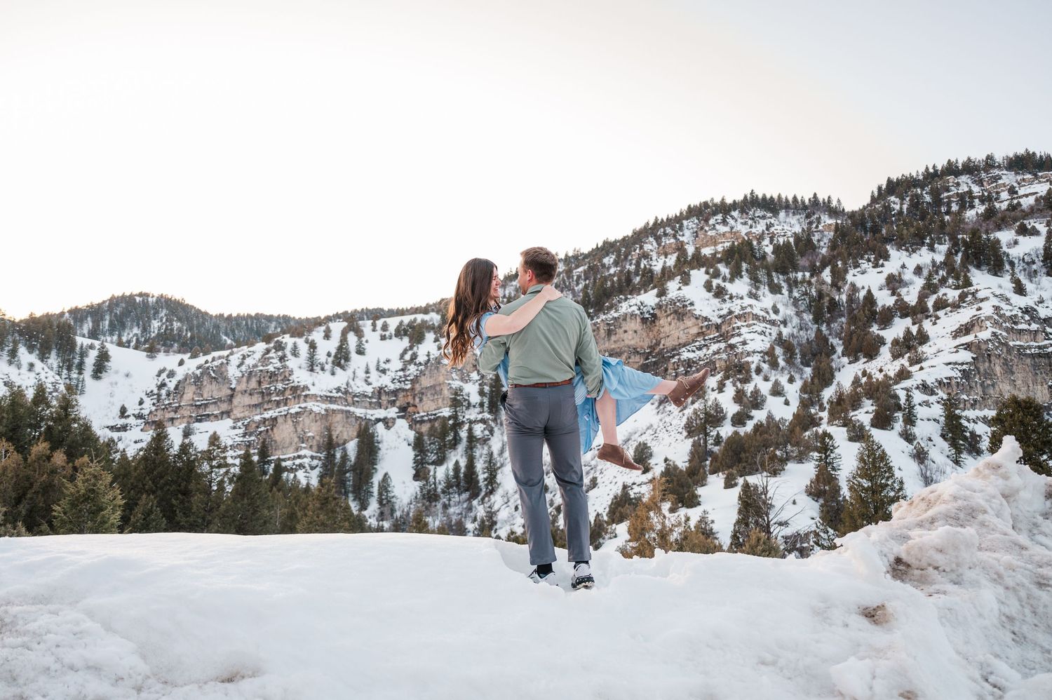 A couple stands on a snowy mountain overlook with evergreen trees and rocky peaks in the background.