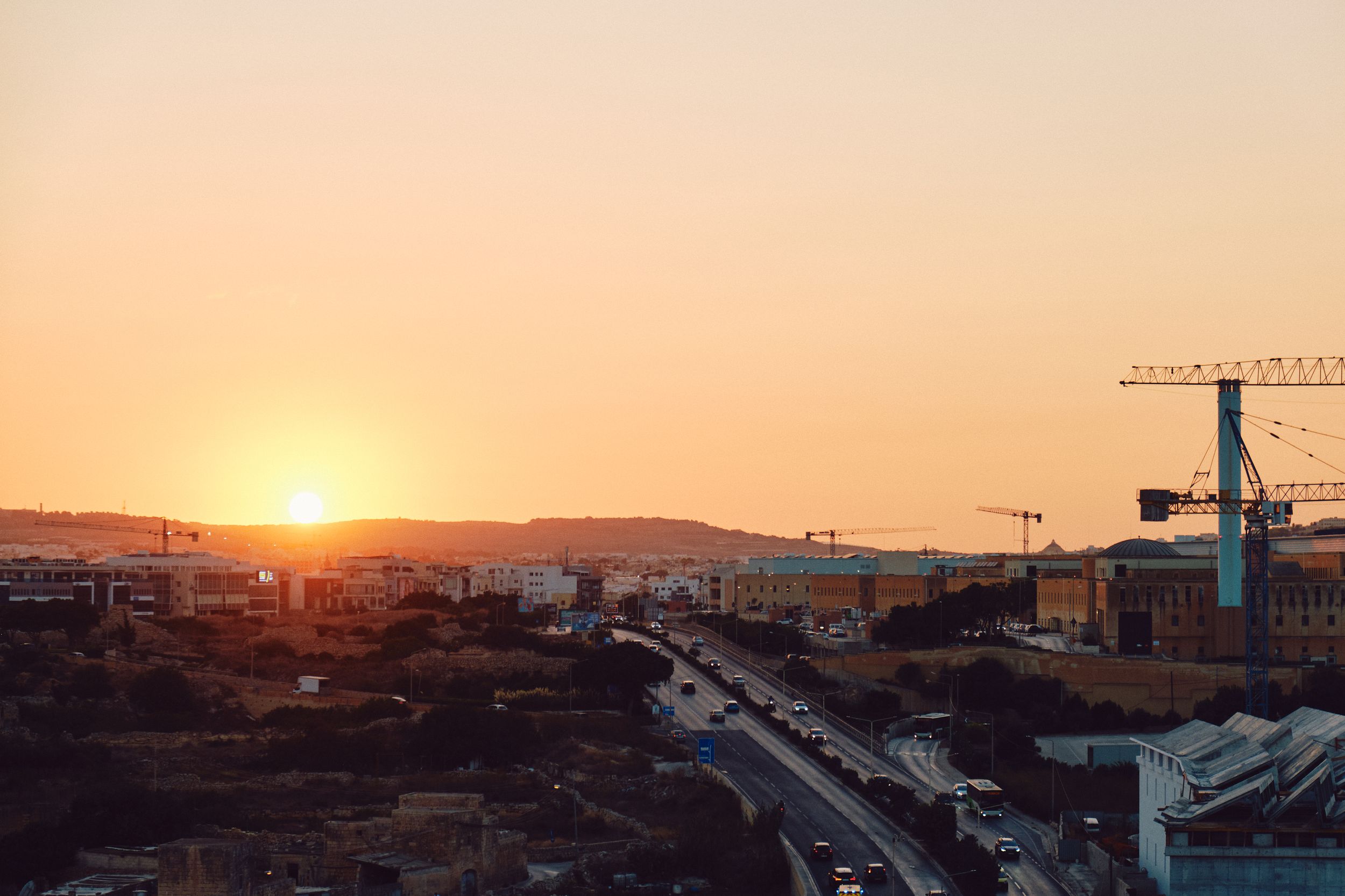 Construction cranes tower over a Mediterranean city during golden sunset.