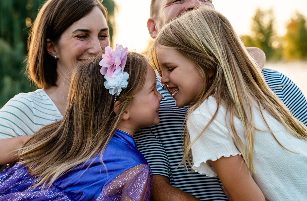 Two young sisters laughing and touching foreheads while parents embrace them warmly outdoors.