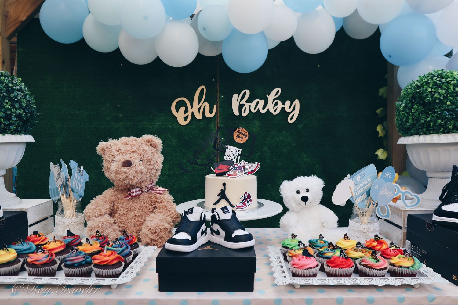 Baby shower dessert table with teddy bears, balloons and cupcakes against green backdrop.