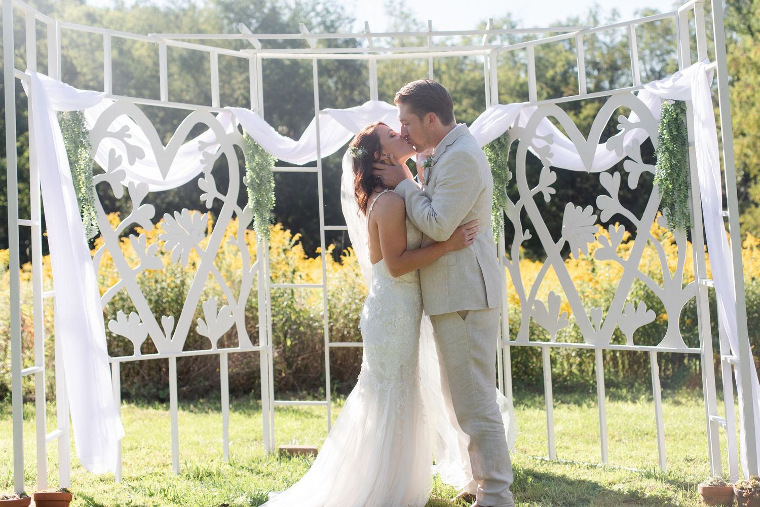 A white fabric-draped wedding arch stands against a blue sky during an outdoor ceremony.