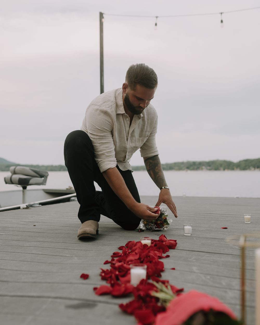 Someone arranging red rose petals in a heart pattern on a wooden deck by the water.