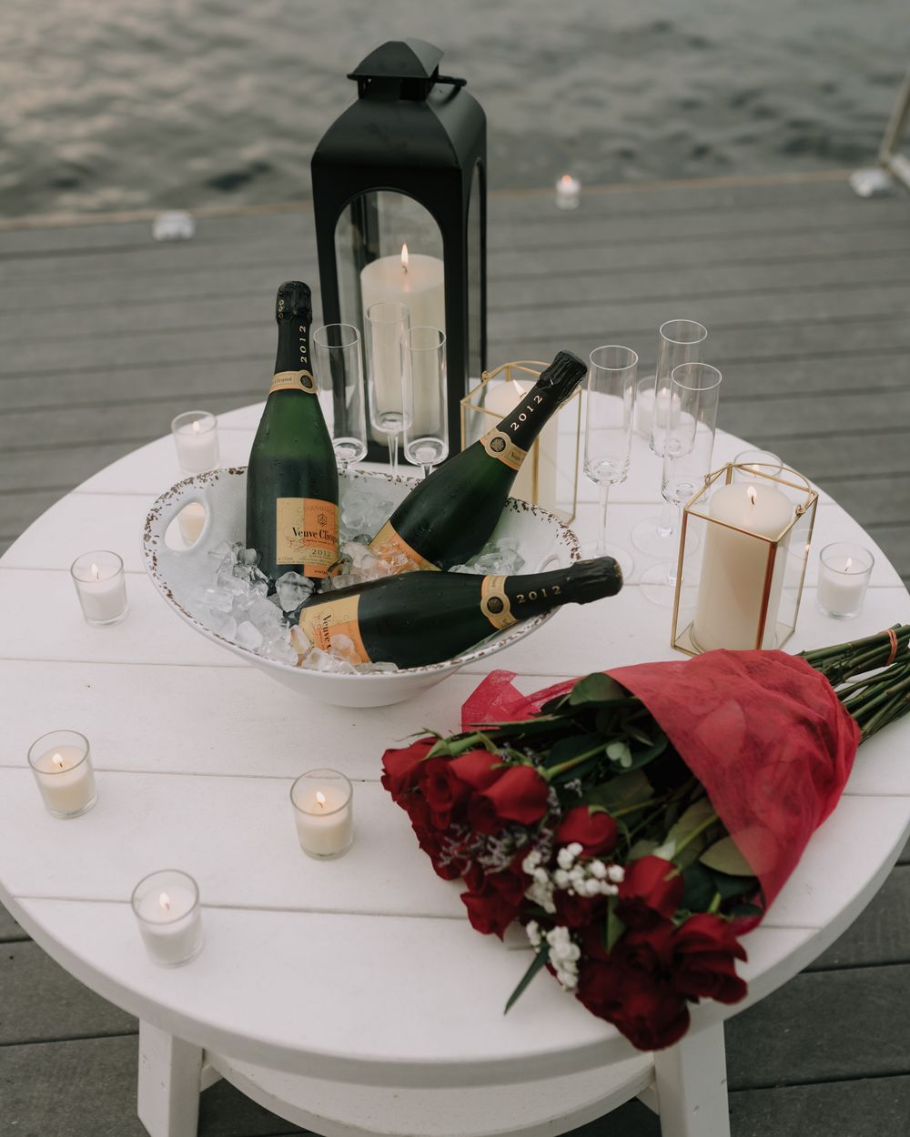 A white round table set with champagne, glasses, and red roses on a wooden deck.
