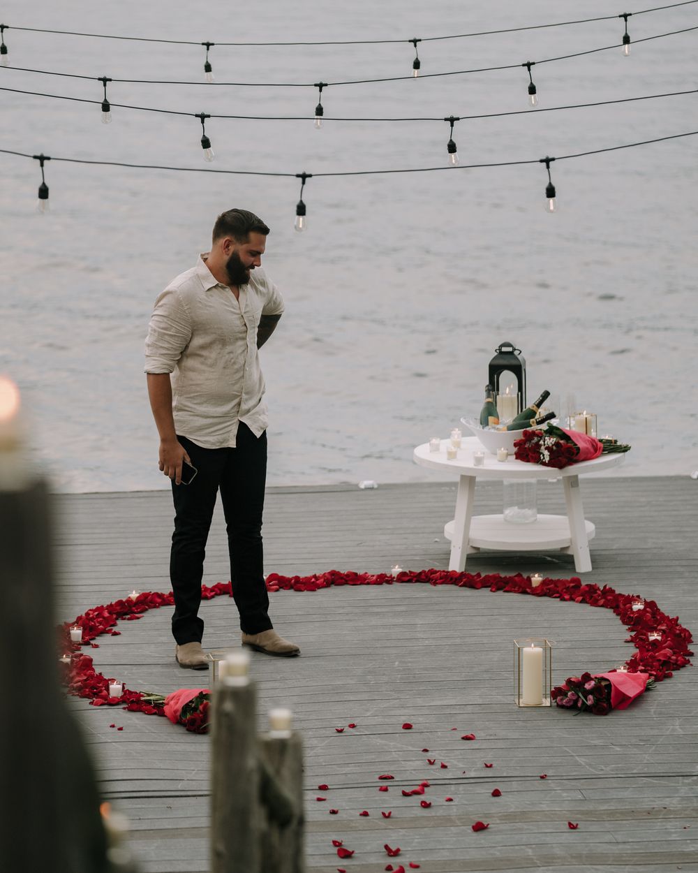 String lights hanging over a deck where red rose petals form a heart pattern with candles.