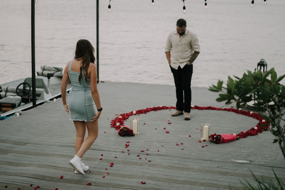 A romantic setup on a wooden deck overlooking water with red rose petals scattered in a heart shape.