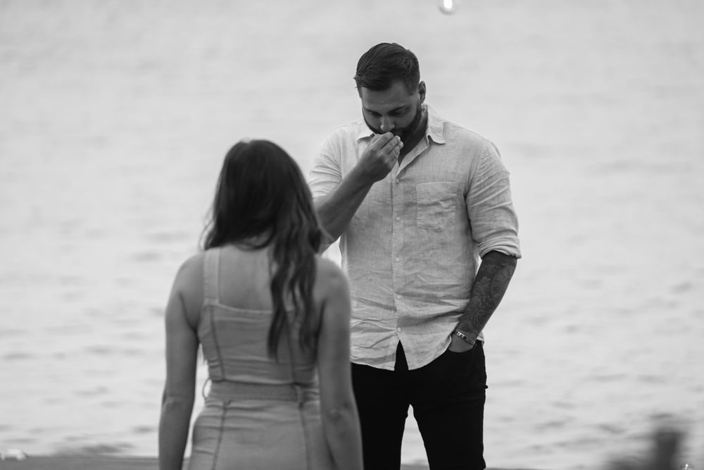 A romantic proposal scene unfolds by the water in black and white.