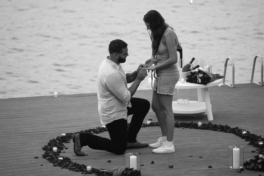 A black and white photograph of a proposal on a dock surrounded by candles and rose petals.