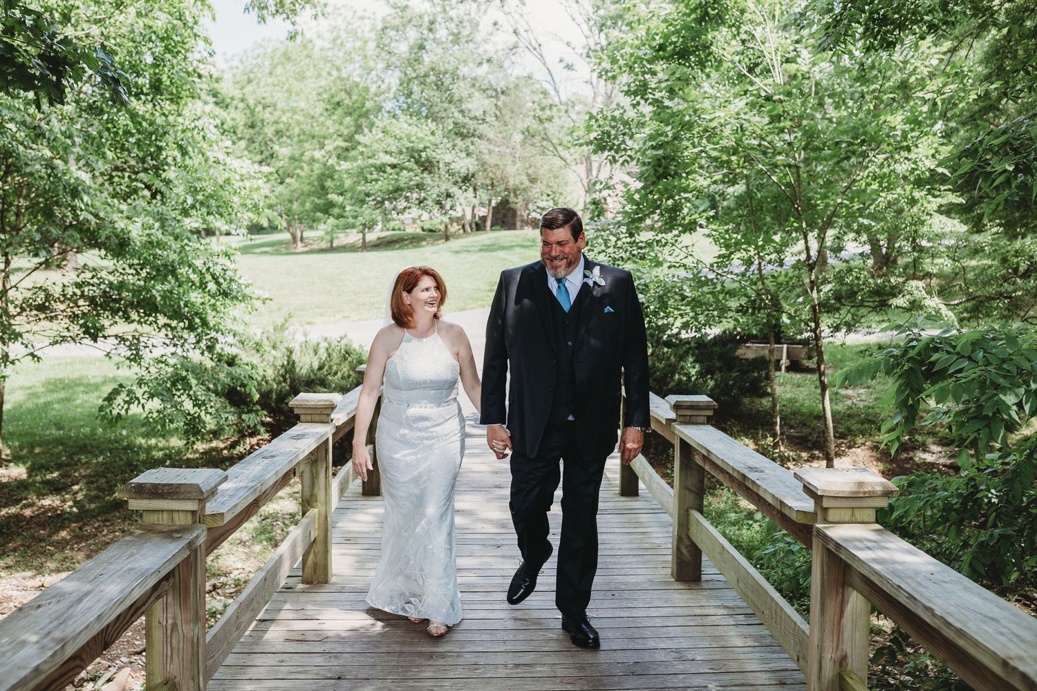 A couple in wedding attire walks along a rustic wooden boardwalk path surrounded by green trees.