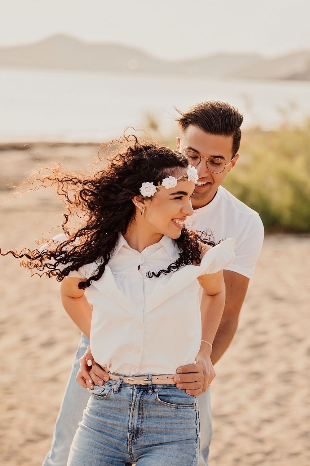 Couple in white tops and jeans sharing playful moment on beach during sunset.