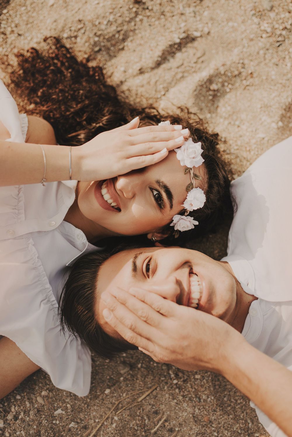 Couple lying on sand playfully covering faces while laughing during photoshoot.