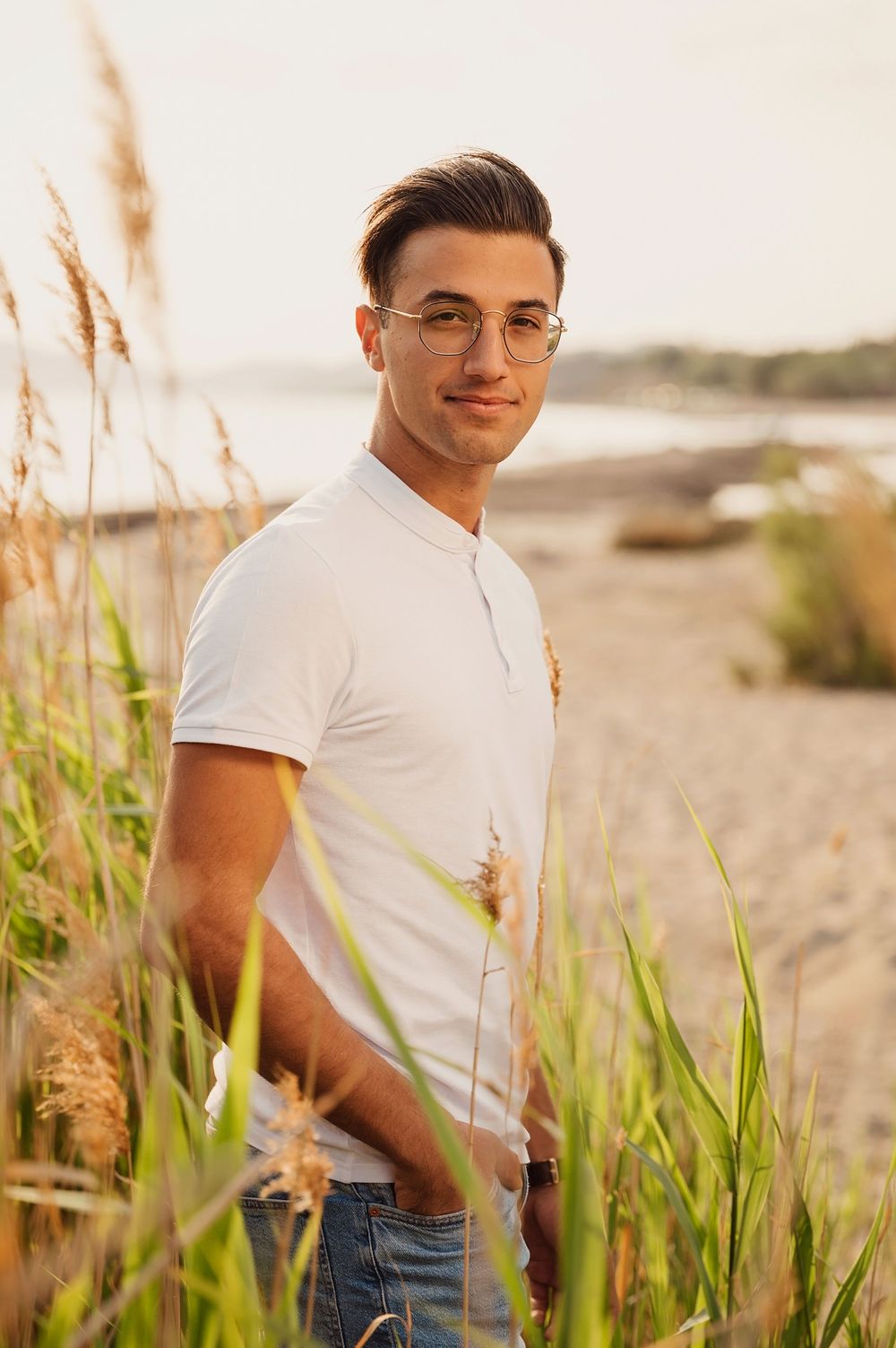 A casual portrait of a young man in white t-shirt standing among beach grass at sunset.