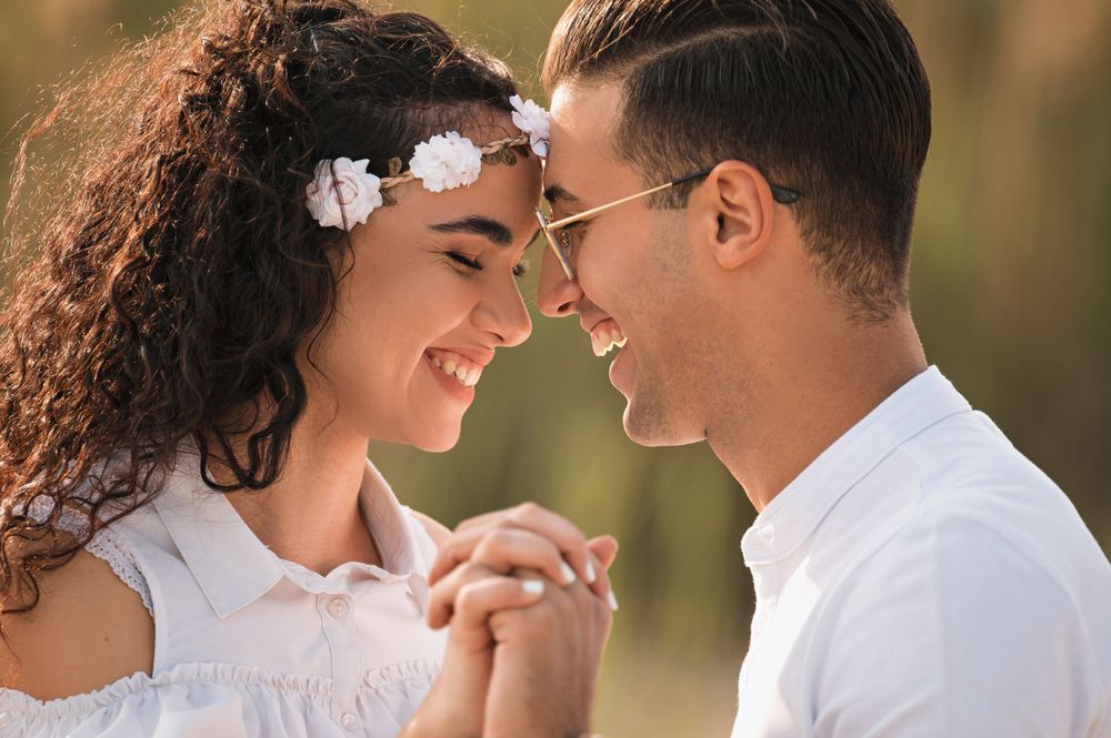 Romantic couple in white outfits smiling at each other during sunset photoshoot.
