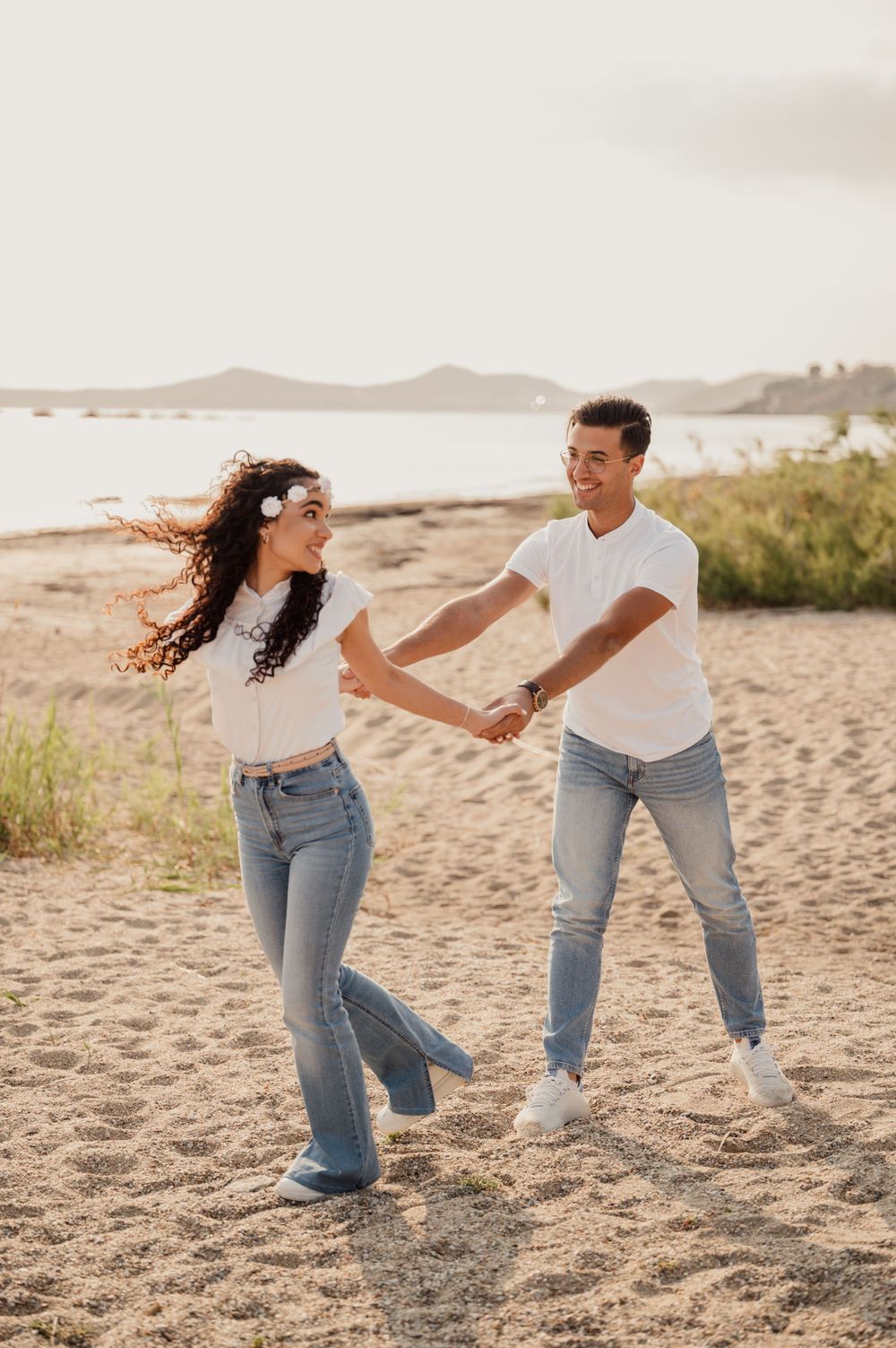 Couple in casual outfits dancing and laughing on beach during sunset.