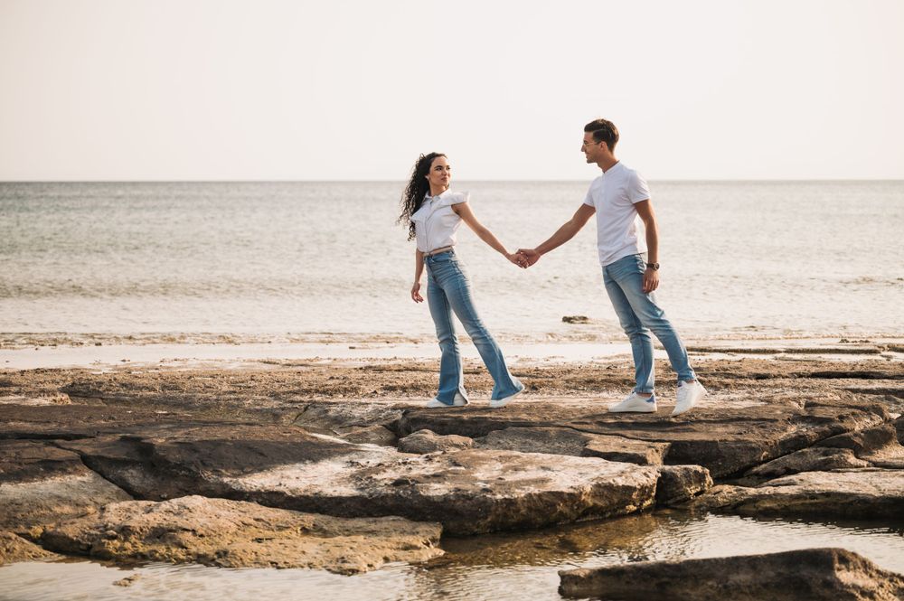 A couple holding hands while walking on rocky beach terrain during sunset.