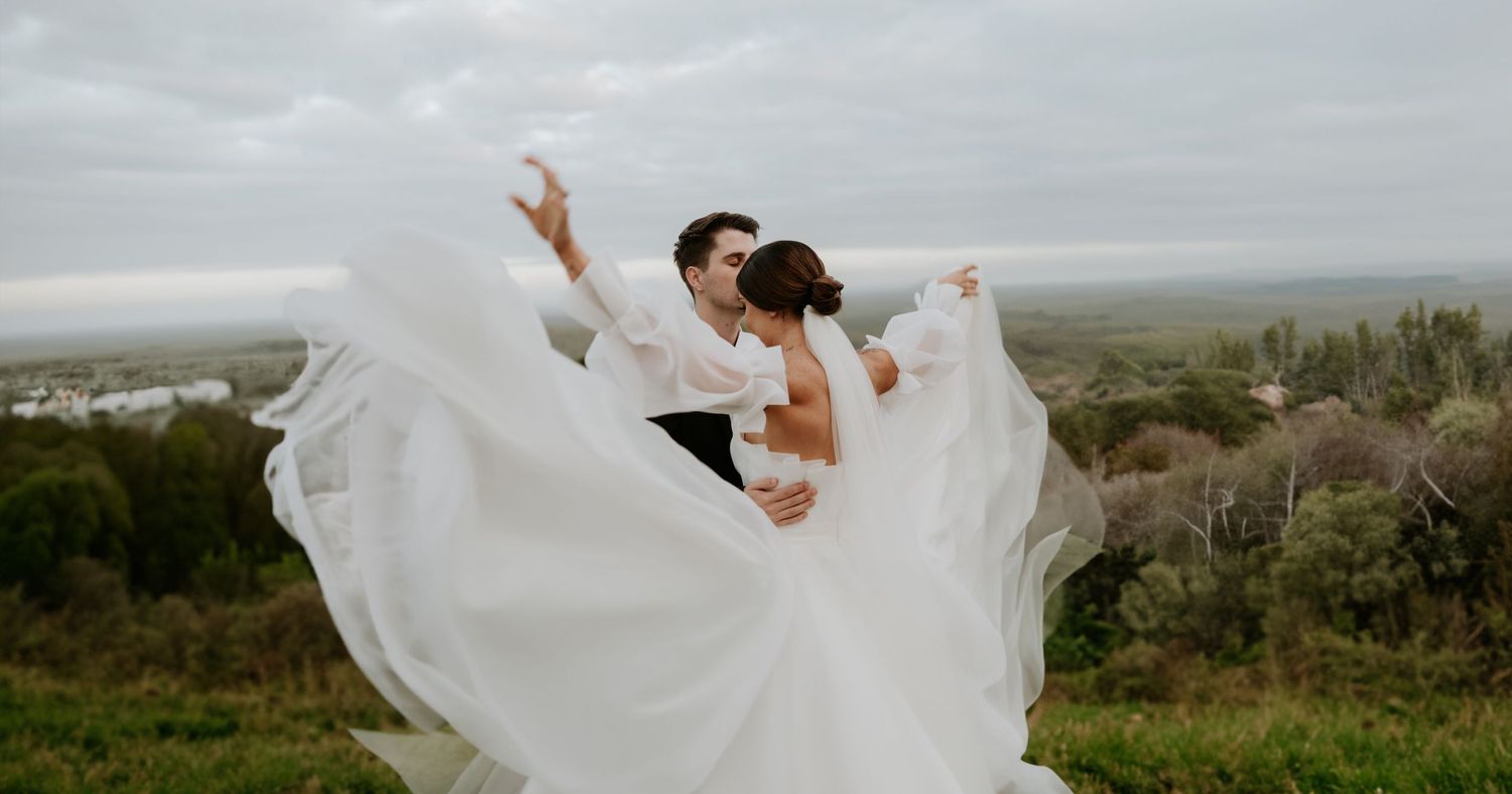 A flowing white wedding dress billows dramatically against a cloudy mountain landscape.
