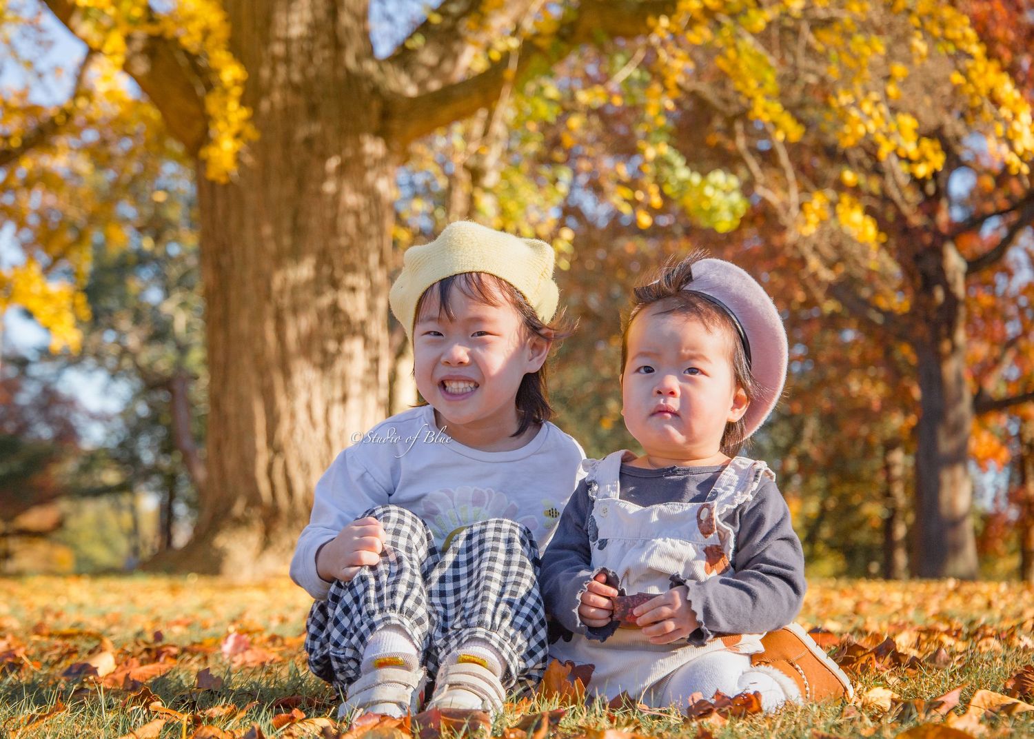 Two young children sit together laughing under golden autumn trees.