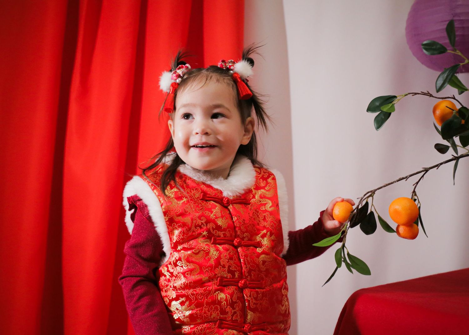 Young child in red traditional Chinese dress smiling by orange tree decorations.