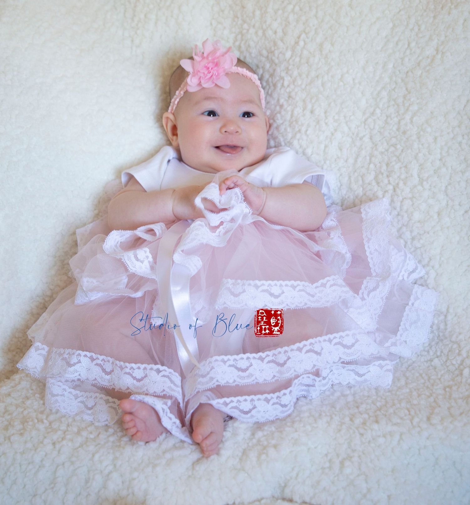 A smiling baby wearing a pink dress and bow headband lies on white textured fabric.