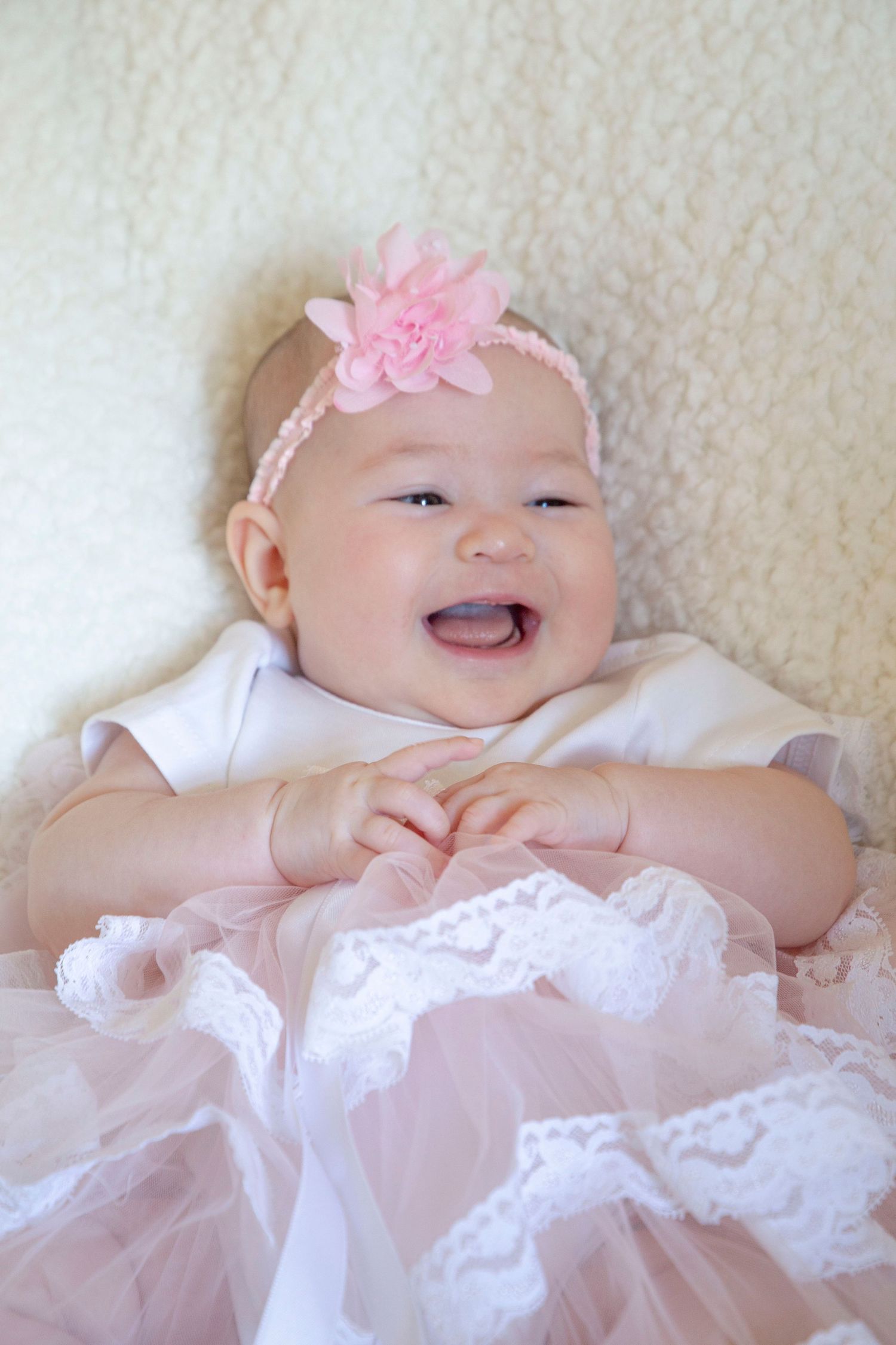 Baby in white dress with pink flower headband lying on white blanket while smiling.