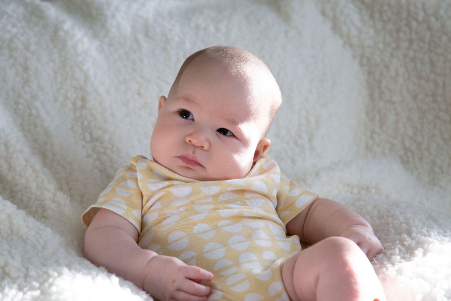 Baby wearing yellow and white striped onesie lying on soft white blanket.