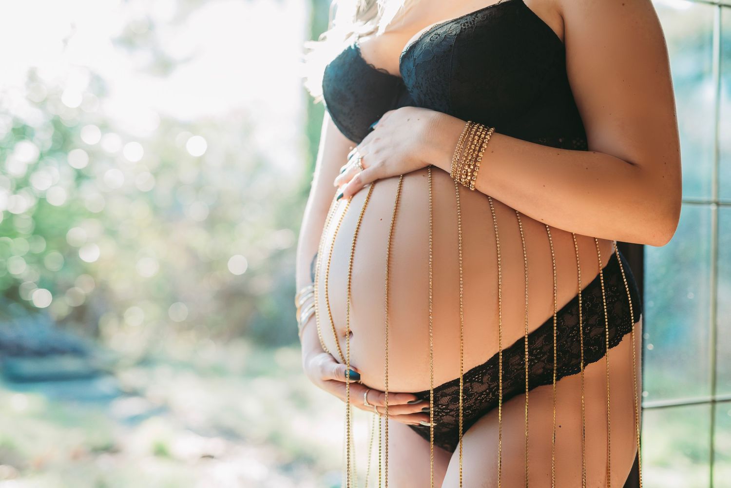 Pregnant woman in black lingerie gently cradles her bare belly in natural sunlight by a window.