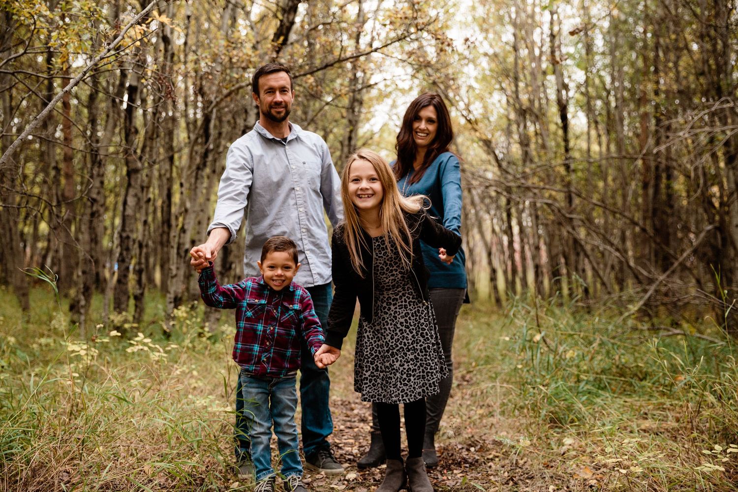 Family of four smiling and holding hands on a forest trail in autumn.