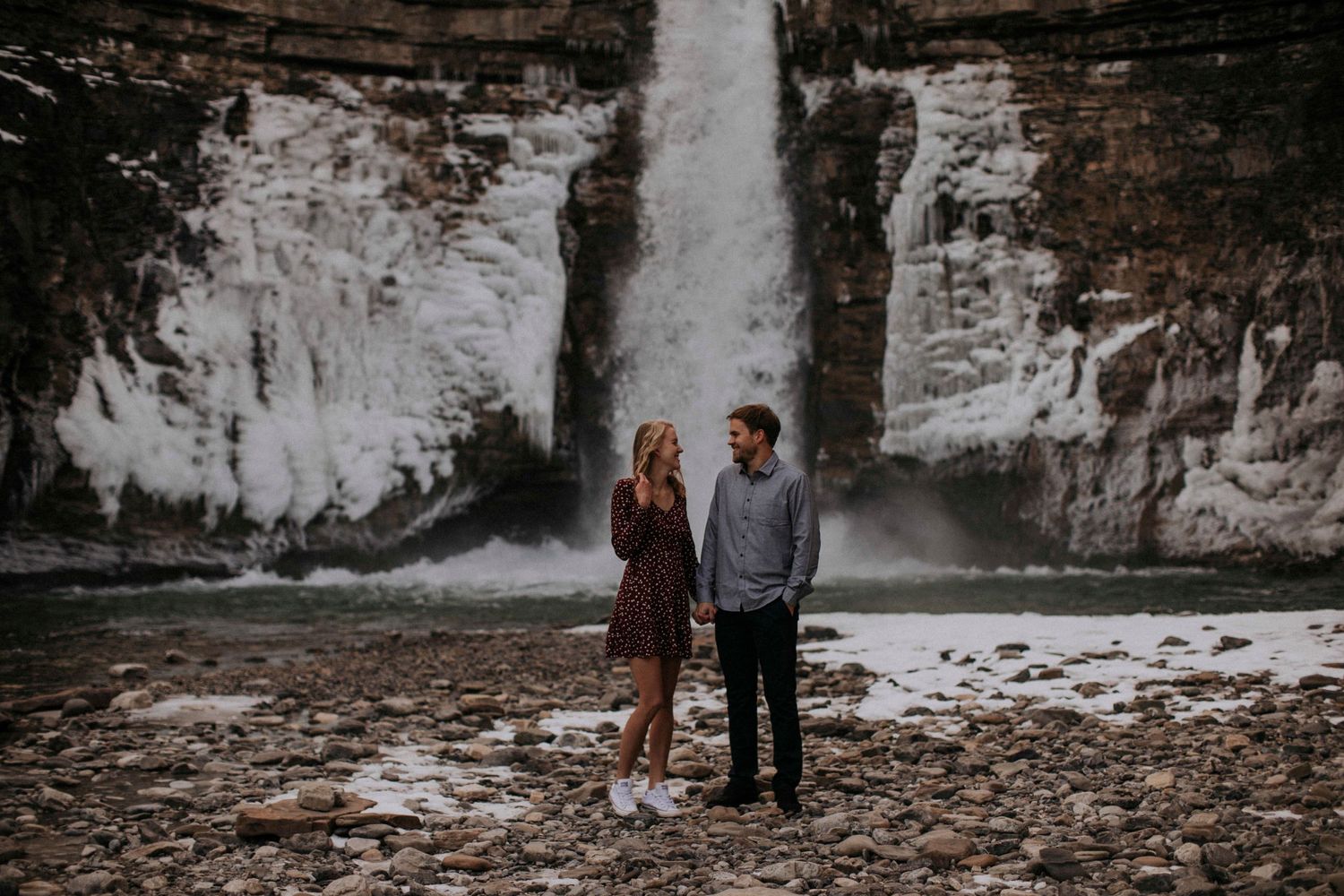 A scenic waterfall engagement photo with a couple standing on rocky terrain beneath towering cliffs.