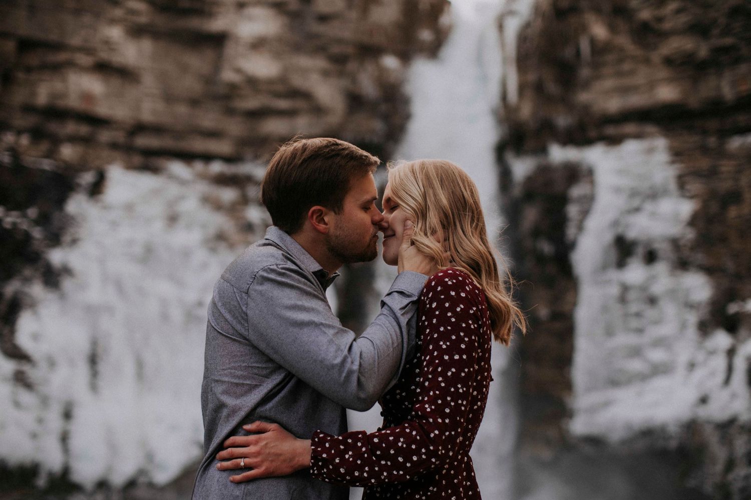 A romantic couple embraces in front of a majestic waterfall on a cloudy day.
