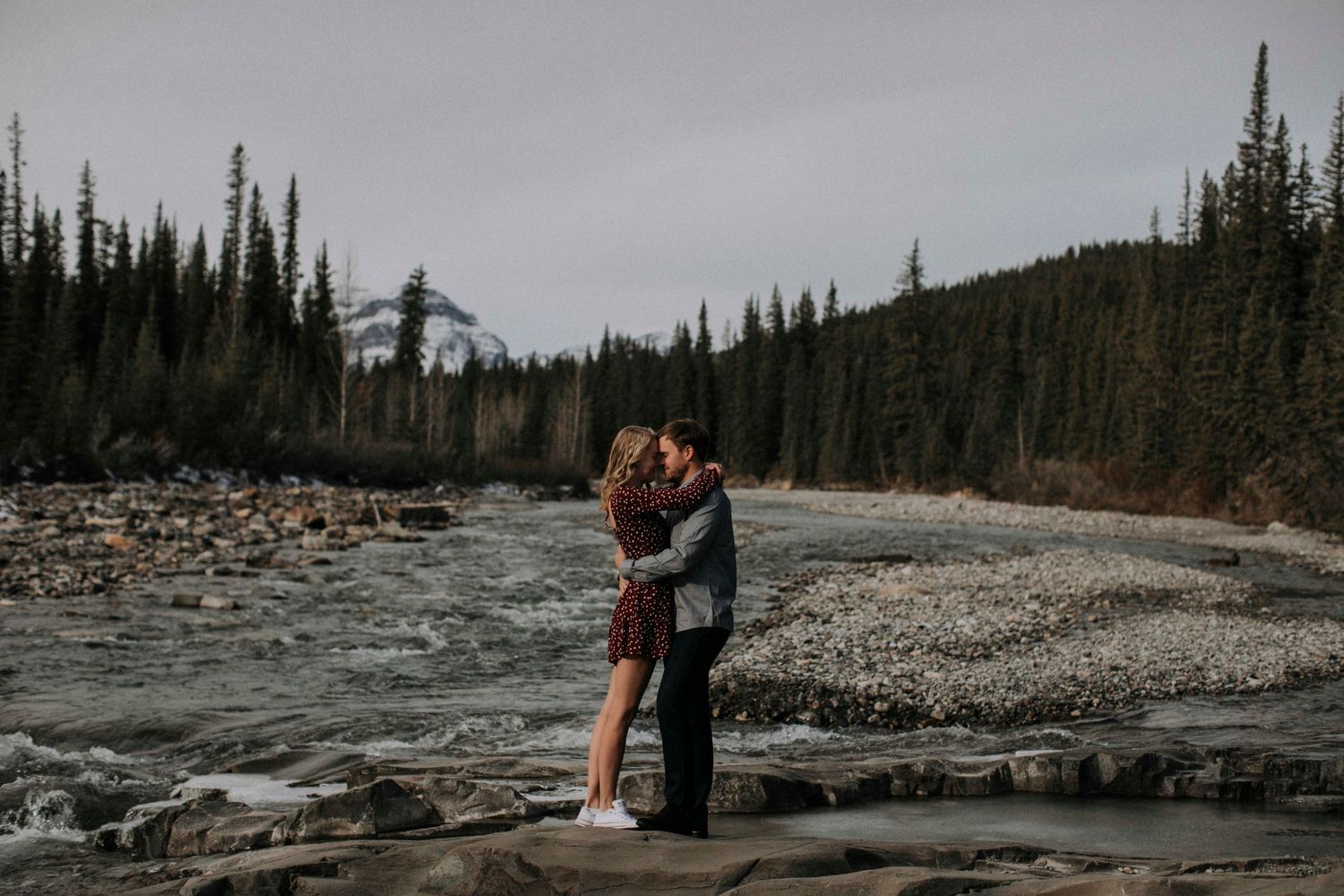 Romantic moment shared beside rushing river with mountain forest landscape.