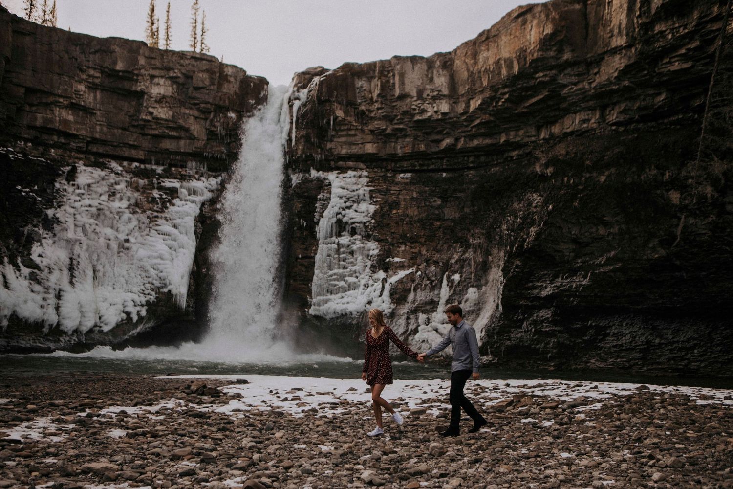 Two people stand holding hands against the backdrop of a dramatic waterfall and rocky cliffs.