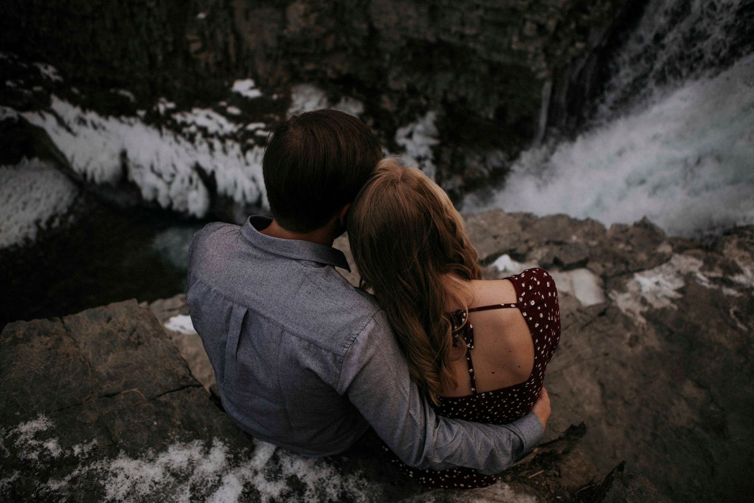 A couple rests together on rocks overlooking a misty waterfall.
